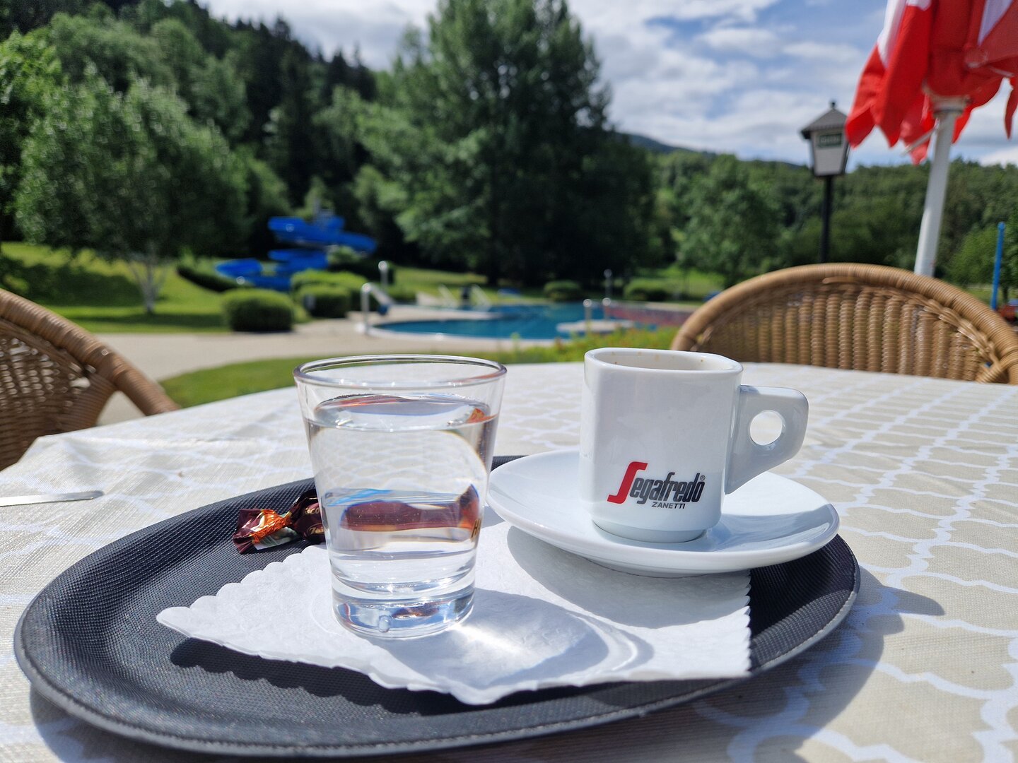 Ein Kaffee mit einem Glas Wasser auf einem Tablet, im Hintergrund schaut man auf das Freibad Gmünd