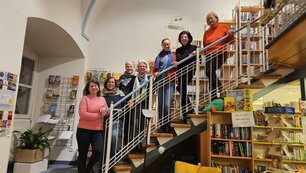 Sieben Damen stehen an einem Stiegengeländer, in dem Raum befinden sich viele Bücherregale