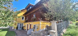 Eine Außenansicht der Ferienwohnung Wieki im Sommer. Der untere Stock des Hauses ist in geld ausgemalen, der obere Stock hat eine Holzverkleidung